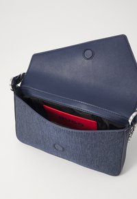 Mørkeblå crossbody-veske i lær og stoff, med kjede-rem og romslig innvendig plass hvor en rød kortholder er synlig.