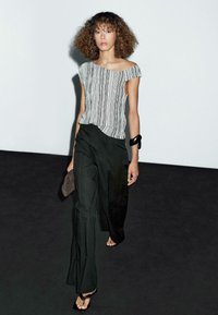 Gestreepte off-shoulder top in zwart en wit, gecombineerd met hoge zwarte broeken. Het model houdt een bruine clutch vast en draagt een zwarte armband.