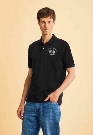 Uomo con polo nera con logo bianco e jeans blu, in piedi davanti a uno sfondo beige uniforme.