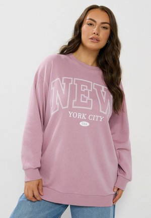 Femme aux longs cheveux bruns portant un sweat-shirt rose oversize avec le texte "New York City USA" et un jean bleu, debout devant un fond blanc.