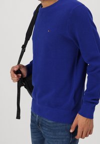 Niebieski sweter o fakturowanej strukturze z okrągłym dekoltem, ściągaczowymi mankietami i dołem. Posiada małe logo na piersi. Noszony z ciemnymi dżinsami i czarną torbą.