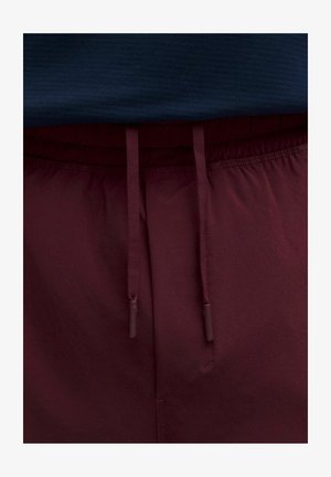 Pantalon bordeaux avec une taille à cordon, présenté avec une chemise bleu marine légèrement rentrée dans la taille.