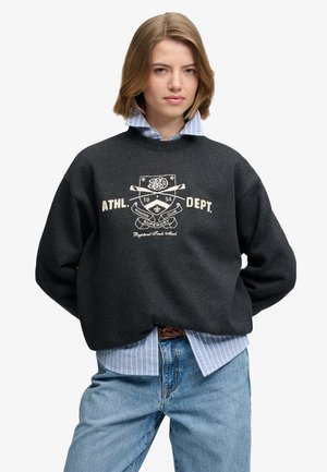 Ung kvinde med skulderlangt blondt hår iført en mørk sweatshirt over en blåstribet skjorte og lyseblå jeans.
