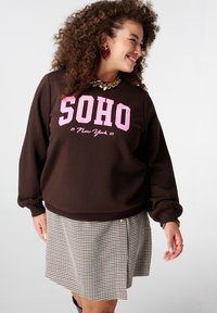 Bruine sweatshirt met roze "SOHO"-print, met lange mouwen en een losse pasvorm, gecombineerd met een geruite rok. Gouden hardware-accenten zichtbaar.