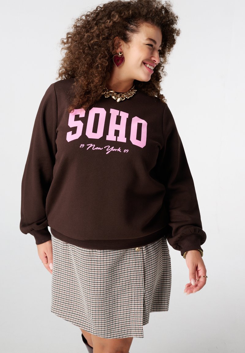 Bruine sweatshirt met roze "SOHO"-print, met lange mouwen en een losse pasvorm, gecombineerd met een geruite rok. Gouden hardware-accenten zichtbaar.