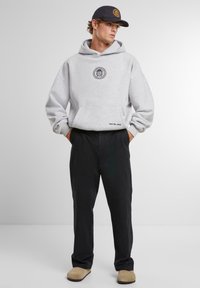 Hanorac gri cu un logo pe piept, pantaloni negri si pantofi slip-on bej, asortați cu o sapcă închisă la culoare cu un logo.