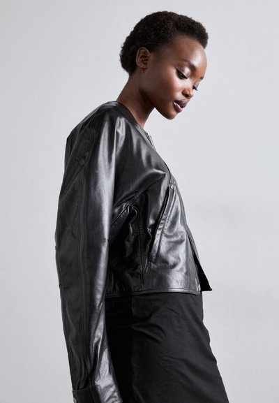 Fekete bőr bombázó dzseki, rövidített kialakítással, oldalsó cipzáras zsebekkel és sima textúrával. Fekete, testhezálló felső fölött viselve.