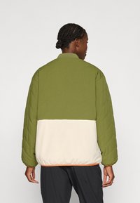 Chaqueta acolchada con una sección superior verde y una sección inferior crema, con un cuello acanalado y una cinturilla elastizada. Patrón texturizado visible.
