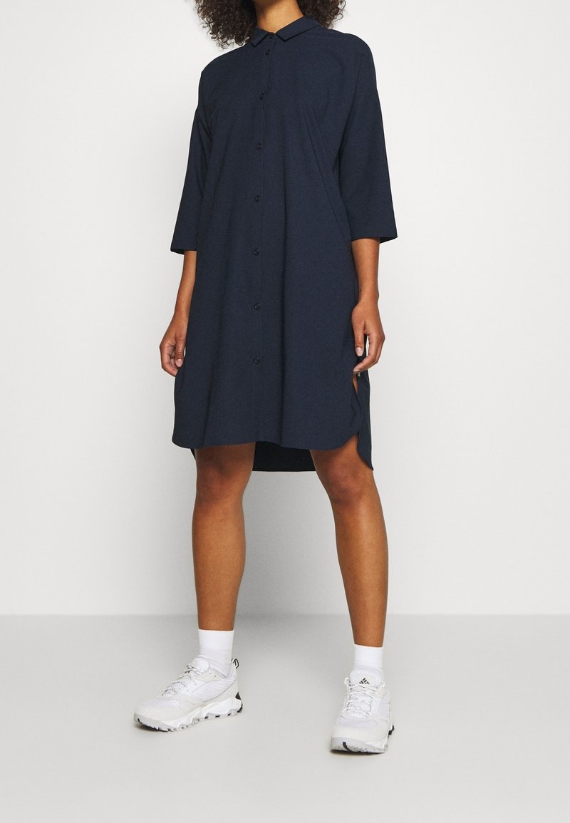Robe bleu marine à col, manches trois-quarts, fentes sur les côtés et coupe décontractée, associée à des chaussures de sport blanches.