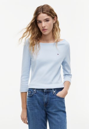 Kobieta o falowanych włosach, ubrana w jasnoniebieską bluzkę z rękawami do łokcia i niebieskie dżinsy, stojąca z jedną ręką w kieszeni na białym tle.