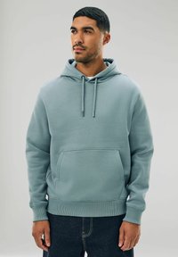 Felpa blu chiaro in tessuto morbido, con tasca anteriore, cappuccio con cordino e polsini a costine. Indossata con jeans scuri.
