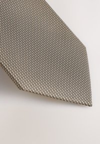Beige Krawatte mit einem strukturierten Muster, das kleine schwarze Quadrate aufweist. Die Spitze ist spitz zulaufend und zeigt eine elegante, glatte Stoffoberfläche.