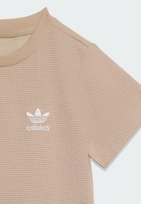Camiseta beige de manga corta con un diseño texturizado de punto waffle. Presenta el logo de Adidas bordado en blanco en el pecho y un cuello acanalado.
