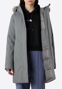 Grå lang frakke med hætte trimmet med pels, der har et quiltet indre for, sidelommer og en afslappet pasform, båret over en sort hoodie.