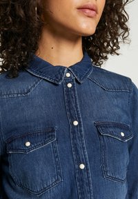 Camicia di denim blu scuro con chiusura frontale a bottoni, colletto e due tasche sul petto. Presenta bottoni a pressione perla e dettagli di cuciture visibili.