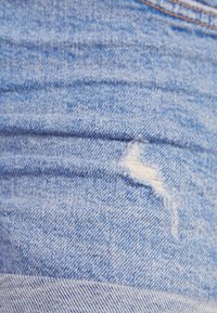 Jeans de denim azul claro con un desgarro deshilachado, mostrando hilos blancos. La tela tiene una superficie texturada con un sutil estriado.