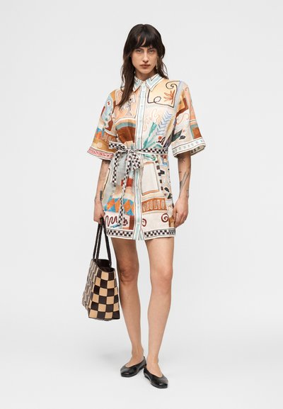 Γυναίκα που φοράει πολύχρωμο φόρεμα με μοτίβα και ζώνη, κρατώντας μια καρό τσάντα ώμου, συνδυασμένο με μαύρα φλατ παπούτσια, σε απλό φόντο.