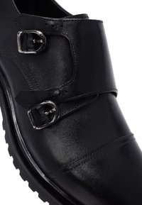 Black leather shoe featuring dual buckles, a sleek design, smooth texture, and a chunky sole. Accented with visible stitching details.