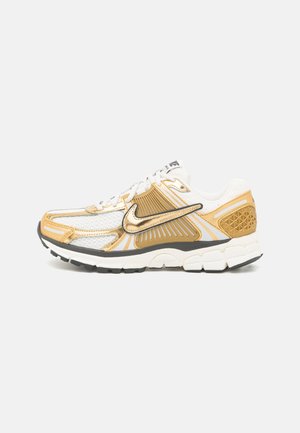 Witte en gouden Nike hardloopschoen met mesh bovenwerk, metalen Swoosh-logo en witte zool met zwarte profiel, zijaanzicht.
