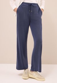 Person klädd i lösa marinblå byxor med dragsko i midjan och beige sneakers, med händerna i fickorna, stående på en ljus golv och bakgrund.