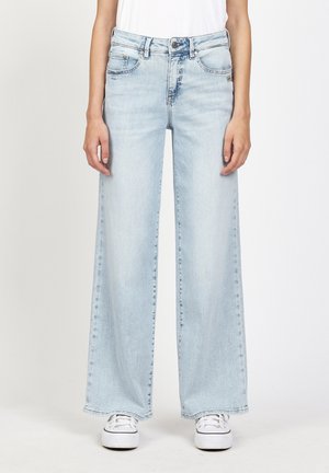 Frau trägt hellblaue Jeans mit weitem Bein und weiße Sneakers, steht vor einem schlichten weißen Hintergrund.