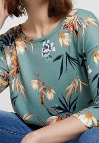 Bluzka w turkusowym kolorze z kwiatowym wzorem, posiadająca rękawy do łokcia, okrągły dekolt oraz miękką fakturę z wielobarwnymi botanicznymi akcentami.
