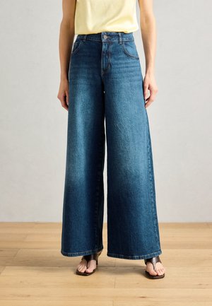 Brede denim jeans in middelblauw, high-waisted met een gladde textuur en zijzakken, gecombineerd met zwarte flip-flops op een houten vloer.