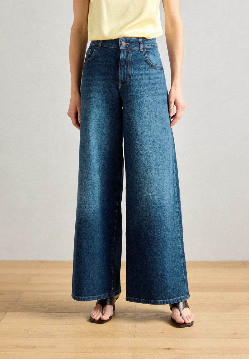 Jeans de denim de perna larga em azul médio, de cintura alta com uma textura suave e bolsos laterais, combinados com chinelos pretos num chão de madeira.