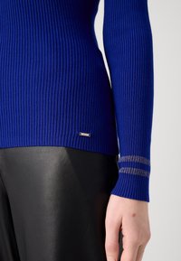 Gros plan d'une main reposant sur le côté, portant un pull côtelé bleu royal avec des rayures métalliques sur le poignet et un pantalon en cuir noir.