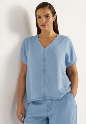 Femme aux cheveux bruns lisses portant un haut bleu clair à manches courtes et col en V, ainsi qu'un pantalon assorti, debout contre un mur blanc.