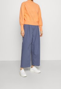 Suéter naranja con cuello redondo y mangas abullonadas, combinado con pantalones azules de corte holgado. Zapatillas blancas completan el atuendo.