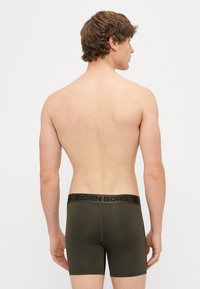Olive zaļas bokseršorti ar melnu jostu, uz kuras ir redzams "BJÖRN BORG" logo. Gluda tekstūra, pieguļoša forma, skatīts no aizmugures.