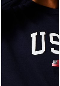 Mörkblå sweatshirt med vitt "US"-tryck och en liten amerikansk flagga som detalj. Tyget verkar mjukt och slätt.
