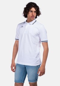 Polo bianco con accenti a righe navy sul colletto e sulle maniche. Presenta una patta con bottoni e un logo sul petto. Indossato con pantaloni shorts in denim.