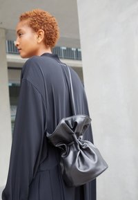 Černá kožená taška s řaseným vrchem, měkkou texturou a dlouhým popruhem, nošená přes dlouhý černý oděv. Minimalistický design s jemnými detaily.