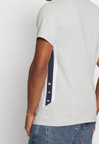 Muž oblečený v světlém šedém tričku s krátkým rukávem, které má černobílé boční pruhy se svislými písmeny "GSRAM", v kombinaci s modrými džínami.