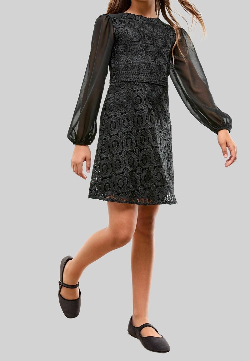 Fille portant une robe noire en dentelle avec des manches longues transparentes et des chaussures Mary Jane noires, marchant avec une jambe légèrement relevée.