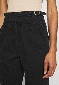 Los pantalones de mezclilla negros presentan una cinturilla triangular única con una hebilla plateada, bolsillos laterales y un ajuste holgado. Tejido texturizado.
