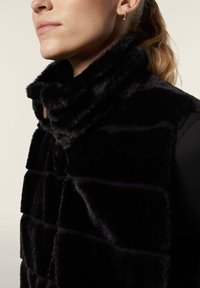 Mustan tekoturkisliivi, jossa on korkea kaulus, tikattu kuosi ja pieni metallilogo, sileä pinta.