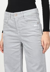 Graue Cordhosen mit vertikaler Rippung, einem Reißverschluss und Knopfverschluss, Fronttaschen und einem aufgenähten Logo an der Seite.