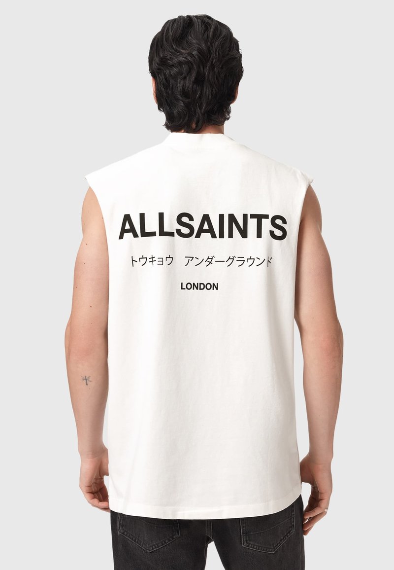 Homme aux cheveux foncés porte un t-shirt blanc sans manches avec le texte "ALLSAINTS," caractères japonais, et "LONDON" au dos, associé à un pantalon noir.