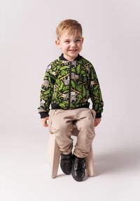 Młody chłopiec uśmiechający się, ubrany w zieloną kurtkę zapinaną na zamek z wzorem leniwca i liści, beżowe spodnie i czarne buty, siedzący na drewnianym stołku.