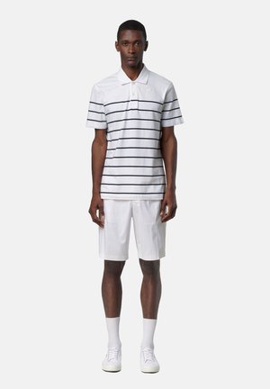 Uomo che indossa una polo bianca con sottili strisce orizzontali nere, pantaloncini bianchi fino al ginocchio, calzini bianchi e scarpe da ginnastica bianche, in piedi dritto.