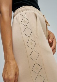 Beige sweatpants met een elastische tailleband, voorzien van diamantenpatronen omlijnd met zwarte en gouden studs langs de zijkant. Zachte textuur.