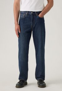 Vaqueros de mezclilla azul oscura con patas rectas, cinco bolsillos, remaches de cobre y cierre de botón. Se llevan con zapatos negros para crear contraste.