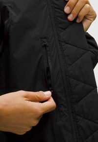 Giubbotto nero trapuntato con tasca laterale con zip, caratterizzato da una texture liscia e un materiale leggero. Mano mostrata mentre afferra il cursore della zip.