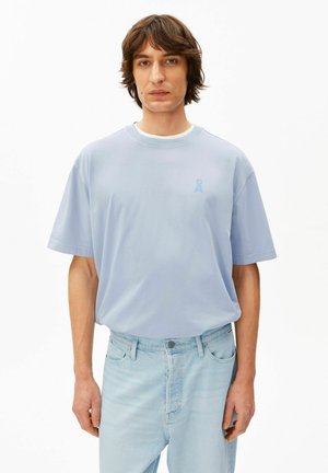 Jonge man met halflang bruin haar, gekleed in een lichtblauw oversized T-shirt en lichtblauwe jeans, tegen een witte achtergrond.