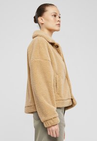 Giacca in pile beige e morbido con colletto arrotondato, chiusura frontale con bottoni e tasche laterali; vestibilità corta con spalle cadenti.