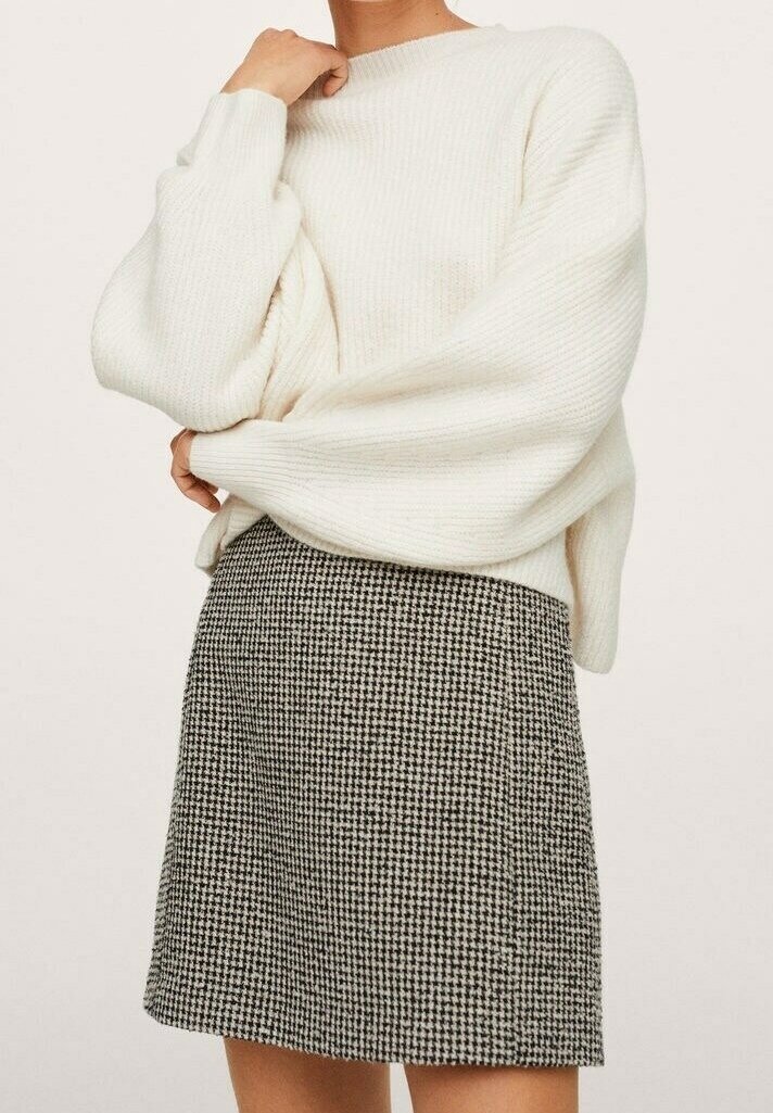 Femme portant un pull en maille blanc oversize et une mini-jupe pied-de-poule noir et blanc, debout, les bras croisés.