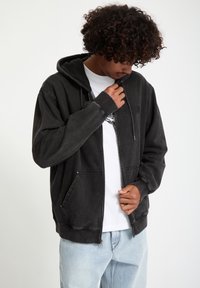Sudadera con cremallera negra de tejido suave y texturizado, con bolsillos laterales y capucha con cordón, sobre una camiseta blanca.